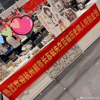 这是一张在动漫展或同人展上的摊位照片，摊位前挂着醒目的红色横幅，内容写着“热烈欢迎杭州市前市长苏轼先生莅临历史同人特别企划”。桌面上摆放着以历史人物苏轼为主角的同人作品，左侧有苏轼的卡牌立牌，并配上了立牌旁的“启禀 集章处”字样。画面融合了现代同人展的亚文化和古代名人穿越的搞笑设定。展台背景有大量苏轼等古代人物相关文创及周边。

这张图采用了“严肃官方横幅”+“历史名人+二次元场景穿越”双关梗，创造出一种极具玩梗氛围的二次元历史同人场景，引发网友调侃和模仿。