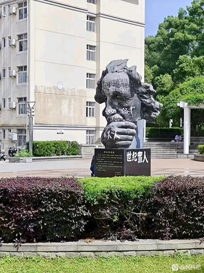 这是一张实拍的公共场景图，原本为阿尔伯特·爱因斯坦的沉思状半身雕像，被人恶意涂写篡改：雕像面部被画上类似口罩的图案和‘必中’字样，手部被涂写‘人人人’，雕像下方的介绍铭牌被替换为写有‘世纪罪人’的标识，原本的人物介绍文字也被更改。场景位于一处带有教学楼和绿植的广场区域，属于对公共艺术作品和名人形象的恶意破坏，带有荒诞的恶搞抹黑性质。