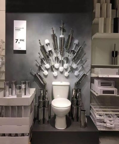 这是一张在宜家门店内拍摄的商品陈列图，画面中心是一个白色马桶，马桶后方的灰色墙面上呈放射状悬挂着多支金属杆的马桶刷，刷头朝向马桶，仿佛将马桶“包围”起来；马桶两侧和周围堆放、摆放着同款马桶刷的金属底座和套装产品，左侧的商品标牌显示这款名为BAREN的马桶刷售价7.99欧元，右侧货架上也陈列着相关卫浴产品。这种夸张的布局制造出趣味视觉效果，能够有效吸引顾客注意力，突出产品的用途。