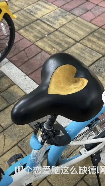 这张图片拍摄了一辆共享单车，焦点在其车座上。车座的表面被抠掉了一大块，而且抠出的形状正好是一个心形，非常不规则又意外浪漫。画面底部配有文字：“哪个恋爱脑这么缺德啊”，调侃了做此事的人把爱心（心型）与“缺德”行为结合起来，形成一种自嘲和幽默的氛围。整体透出网络上常见的恶搞、吐槽文化。

本图包含双关和暗示：车座上的心形缺口既有“爱心”之意，同时“抠心”与“缺德”形成谐音、双关效果。
