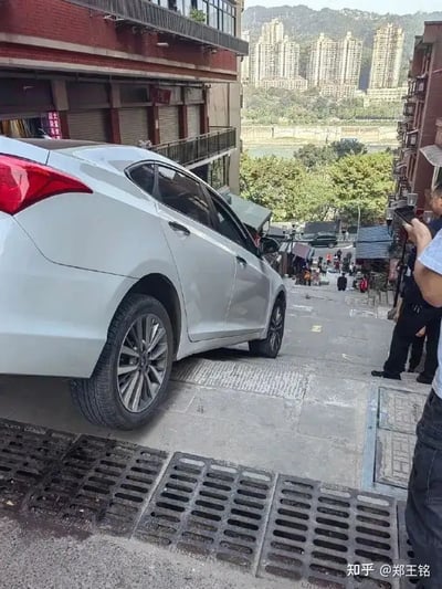 这张图片展示了一辆白色轿车停在城市陡峭阶梯的上方前端，看起来像是误将台阶当成了马路，造成车头悬空，仿佛随时都会下滑。旁边有几位行人围观和拍照。背景为典型的西南城市地形，远处高楼林立。画面引发网友调侃：『重庆坡多路怪，导航都懵了』，常作为城市奇闻、搞笑乌龙或路况调侃图流传。没有发现明显双关或谐音梗，但重庆城市特殊的地形反差是笑点。