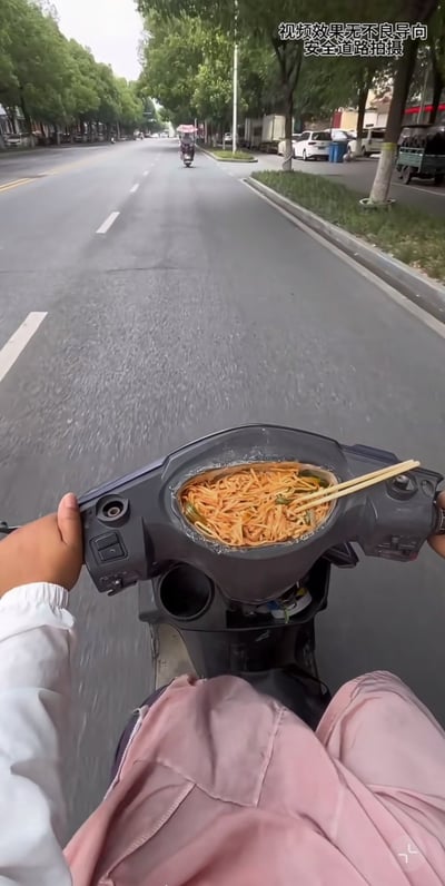 这张图片拍摄于城市道路上，视角为一个人骑电动车的第一视角。最吸引眼球的是电动车的仪表盘位置正好放着一碗面，筷子直接插在碗里，骑手一只手扶着把手，一只手疑似准备夹面，透露一种“边骑车边吃面”的极致离谱感。画面上方有文字“视频效果无不良导向 安全道路拍摄”，说明是特意拍摄的效果，并非生活常态。

有很强的反差和荒诞意味，既有生活气息，又带有一定的搞笑和危险暗示。