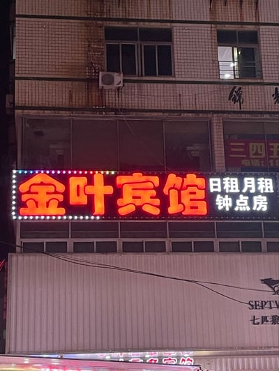 这是一张夜晚拍摄的临街建筑实拍图，画面核心是一块安装在建筑外的发光招牌，招牌左侧是四个醒目的橙色亮灯大字“金叶宾馆”，右侧是白色亮灯标注的经营内容“日租月租 钟点房”，招牌边缘环绕装饰有白色和彩色的小灯珠。招牌上方是贴有米白色小瓷砖的多层建筑，能看到窗户和外挂空调外机，招牌下方是带有七匹狼品牌标识的商铺外立面，整体是国内街头常见的中小型住宿商户的招牌场景。
