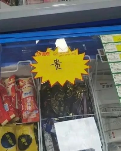 这张实拍图展示了商家存放冰棍的卧式冰柜内部，冰柜被分隔为多个区域摆放不同包装的冰棍产品，在其中一个摆放黑色包装冰棍的区域上方，贴有一个黄色锯齿边的爆炸形提示贴，贴纸上没有标注具体价格，仅用黑色字迹手写了一个醒目的“贵”字，直白提示该区域的冰棍售价较高，冰柜右侧边缘还可见贴有商品价格的标签栏。