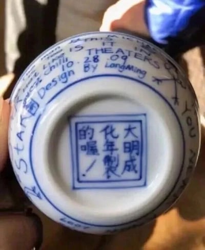 图片展示了一件青花瓷的底部特写，中心为方形印章式底款，内有蓝色文字“大明成化年製的喔！”，其中“的喔！”为口语化后缀，与传统官窑底款的严肃风格形成反差。底款周围环绕着英文文字“STAR THEATERS”“28.09”“Design By Langming”等现代元素，整体呈现出对传统古董瓷的幽默改编，通过古今文字混搭制造搞笑效果。