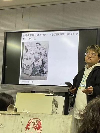 美术课堂上，一位教师站在投影屏幕前讲解模考试卷。屏幕上展示着一张被标记为“零分”的素描作品，标题为《收到快递的一瞬间》，要求绘制“一男一女”的场景。画中男性角色正从纸箱中抱起女性，动作略显夸张。教师手持手机，面前摆放着学生的画板，似乎在点评这张“奇葩”试卷，课堂氛围带有幽默与无奈的气息。