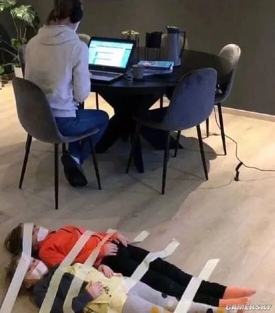 这张图片是一张充满黑色幽默的原始生活照。画面中，一位穿着灰色连帽衫、头戴耳机的成年人背对着镜头，坐在餐桌前专注操作笔记本电脑，显然正在进行居家办公或线上学习。在她前方的浅色地板上，有两个戴着口罩的孩子被白色胶带交叉缠绕着“固定”在地面，安静地躺着。这个夸张且戏谑的行为，实则是家长为了避免孩子吵闹干扰工作而想出的搞笑办法，生动地展现了职场家长在居家办公时，需要同时兼顾工作与照顾孩子的窘迫与无奈，用荒诞的方式放大了这种生活困境，引发同类人群的共鸣。