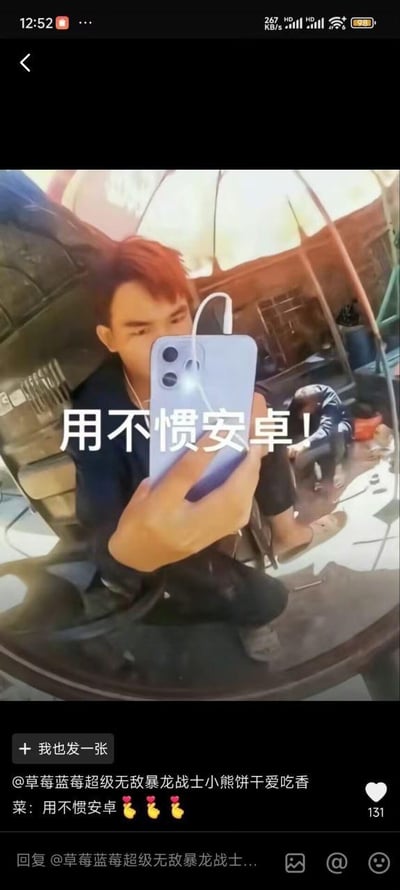 这是一张来自社交平台的搞笑反差梗图，画面中一名红头发男子蹲在类似汽修车间的环境里，对着镜子用一部带有有线耳机的iPhone手机自拍，画面中间用白色大字标注“用不惯安卓！”，形成强烈的反差感，用反讽的方式调侃那些明明使用苹果手机却声称不习惯安卓系统的行为。截图下方显示发布者账号为@草莓蓝莓超级无敌暴龙战士小熊饼干爱吃香菜，其配文同样是“用不惯安卓”并附带三个比心表情，该内容获得了131个点赞，属于网络上流行的趣味吐槽类内容。