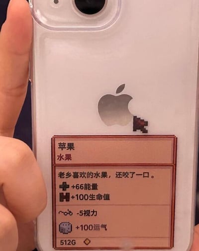 这张图片是一部带有苹果（Apple）Logo的手机，其手机壳上贴着仿照像素风游戏道具卡片的贴纸，把苹果Logo当做游戏道具“苹果”，描述栏还写了各种“属性”和“加成”。贴纸底部还特别写明“还咬了一口”，暗指苹果Logo缺了一块。同时，属性里有体能、生命值、视力（负面效果）以及运气等，非常搞笑有梗。

**谐音/双关：**
- 苹果（水果）与Apple品牌Logo双关，并结合苹果标志的“被咬一口”梗。
- 游戏属性表述+生活真实体验调侃（比如吃苹果补充体力、运气等）完全娱乐化。
