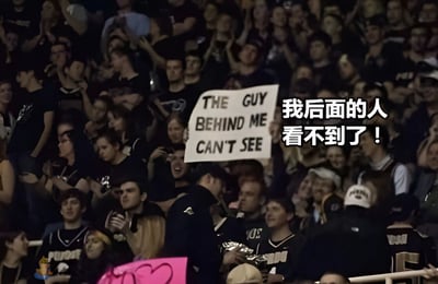 一张体育场观众席的照片，一名观众举着写有'THE GUY BEHIND ME CAN'T SEE'的白色标语牌，图片右侧叠加了中文翻译'我后面的人看不到了！'。照片捕捉了体育赛事中观众席的热闹场景，背景中可以看到许多站立欢呼的观众，部分人身穿带有'PURDUE'字样的服装，暗示这可能是在美国普渡大学的体育赛事现场。前景中有一位穿着制服的安保人员正在巡视。