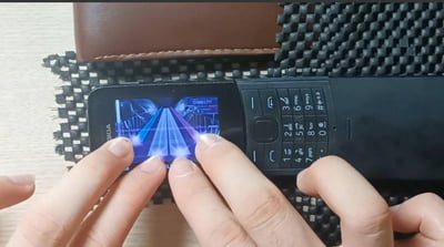 这张原始拍摄的横图展示了一双手正专注地操作一台诺基亚按键功能机，手机屏幕上运行着一款类似《节奏大师》的下落式按键音乐游戏，屏幕中显示着蓝色的轨道判定线、即将下落的按键提示，还有分数“01985711”。双手的手指正按在手机下方对应位置，模拟游戏的按键操作。手机放置在黑色网格状的垫子上，背景还有一个棕色皮质物品，整体画面充满复古怀旧感，唤起人们对功能机时代用按键机玩小游戏的回忆。