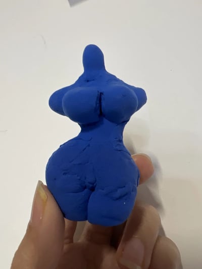 一只手握着一个蓝色的泥塑作品，该作品呈现出抽象的女性人体形态，具有夸张的胸部和臀部曲线，整体造型简洁粗犷，表面可见明显的手工捏制痕迹和指纹纹理。背景为纯白色，突出展示泥塑主体。