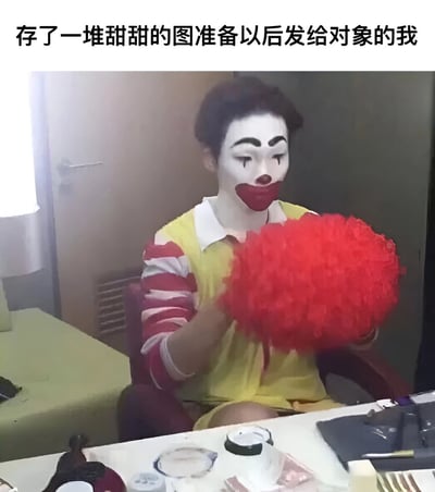图片上方有文字'存了一堆甜甜的图准备以后发给对象的我'，下方是一位化着小丑妆（白色脸妆、红色圆鼻子、黑色粗眉毛）的人，穿着类似麦当劳叔叔的黄红条纹上衣，坐在桌子前，手里拿着一个巨大的红色绒球。整体营造出一种自嘲幽默的氛围，暗示自己像个小丑一样存了很多甜蜜图片却可能没有对象可以发送。