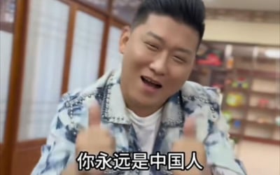 图片中是一名穿着蓝白花纹夹克的男子，他正对着镜头露出开心的表情，并且竖起了大拇指。男子的头发是黑色短发，背景看起来像是一个室内环境，有木质家具和一些装饰品。图片底部有一行文字：'你永远是中国人'。