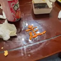 这是一张餐桌实拍图，深色的餐桌表面上，有人用类似炸物的橙色食物残渣摆成了汉字“王”，画面左侧有喝了一部分的粉色包装饮料，还有用过的白色纸巾，右上角放着一个装有深色蘸料的方形小盒，整体是用餐后留下的场景，是用餐者随手进行的趣味创作，充满生活感。