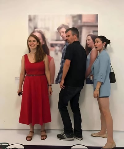 图中展示了三个人在一幅大型照片前的有趣互动场景。左边的红衣女子面带微笑，中间的黑衣男子正专注地看着照片，而右边的蓝衣女子则露出惊讶的表情。照片背景似乎是一个城市街道场景，整体氛围像是在艺术展览或画廊中。