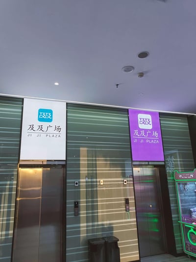 这是一张室内实拍图，展示了及及广场的两部电梯区域。左侧电梯上方是白底蓝标的“及及广场 JI JI PLAZA”标识，右侧电梯上方是紫底白标的同款标识，电梯为金属质感的轿厢门。电梯旁的墙面上设有电梯呼叫按钮，右侧还有一台带有“THE Bear haunt”字样的抓娃娃机，天花板上可见消防报警器、通风口和嵌入式顶灯，整体是商场内的日常电梯场景，无谐音梗、双关或暗示内容。