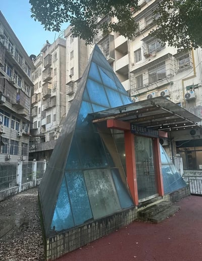 这张实拍图展示了一处建在老旧居民楼之间的小型临街商铺，该建筑将三种不同文化的标志性建筑元素进行了融合：整体为四棱锥的蓝色玻璃结构，外形参考了埃及金字塔的形态，玻璃金字塔的设计又和法国卢浮宫的入口金字塔高度相似，而商铺的入口采用了红色立柱搭配平顶雨棚的设计，外形酷似日本的鸟居。建筑表面有明显的水渍、灰尘等使用痕迹，商铺门口悬挂的招牌显示其为钻机配件材料经营部，周边是布满防盗网的老式居民住宅，地面一侧有堆积的落叶，另一侧是红色塑胶步道，整体风格混搭猎奇。