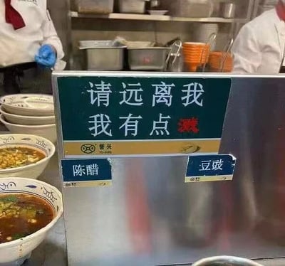 这是一张原始拍摄的静态照片，拍摄场景为中式面馆的出餐区域。画面中心是一个不锈钢材质的调料台，台面上设有两个蓝色小标识牌，分别标注“陈醋”和“豆豉”，指示调料品类。调料台上方的绿色标牌上写着“请远离我 我有点溅”的标语，这里运用了谐音双关的手法，“溅”一方面指调料台的汤汁、调料容易溅出，提醒顾客保持距离，避免弄脏衣物；另一方面谐音“贱”，用诙谐幽默的语气来表达，让生硬的提示变得有趣。画面左侧摆放着两碗已经盛好的面食，面条汤汁浓郁，看起来是店内的餐品。背景中可以看到两位身着白色后厨工作服的工作人员，其中一位戴着蓝色手套，似乎正在进行后厨操作，整体场景真实还原了面馆的日常运营环境，充满生活气息。