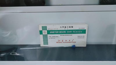这是一张实验室场景下的原始实拍图，画面主体是北京化工厂生产的六甲基二硅醚核磁共振分析试剂包装盒，盒前摆放着一支装有该试剂的安瓿瓶。包装盒上清晰标注了试剂的中文名称、英文名称、分子式、分子量、规格（10×0.5毫升）、批号820522，同时注明该试剂为核磁参比试剂且核磁试验合格，是专门用于核磁共振分析的化学试剂，整体呈现出专业的实验室用品特征。