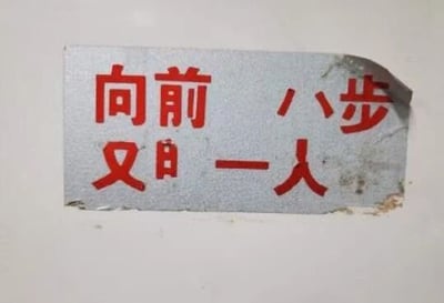 这是一张拍摄于生活中的静态照片，展示了一张贴在白色墙面上的银色底红色字标语贴纸。贴纸因右上角和底部的破损，原本的提示文字“向前一步，又多一人”被破坏成了看起来像是“向前八步，又日一人”的内容，意外形成了低俗又搞笑的歧义效果，属于物品破损偶然造成的趣味文字场景。