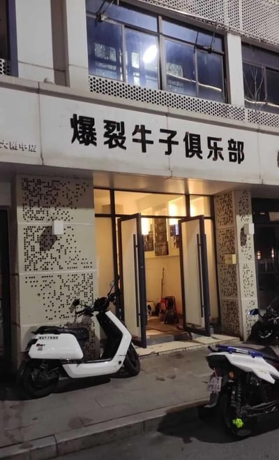 这是一张夜晚街头的店铺实拍图，画面主体是一家名为「爆裂牛子俱乐部」的店铺，白色招牌上的黑色大字十分醒目，店铺门敞开着，能看到店内摆放着清洁工具等物品；店外的人行道上停放着一辆白色电动车、一辆带蓝色装饰的电动车和一辆摩托车。
**低俗梗说明**：「牛子」在中文网络语境中是低俗俚语，该店铺使用这个词作为店名，属于利用猎奇低俗词汇来吸引眼球，制造趣味感。