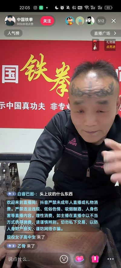 这是抖音平台的一场直播截图，主播「中国铁拳」是一位额头带有龙形纹身的中年男性，他坐在印有「国铁拳」「示中国真功夫 非专业」字样的红色背景板前进行直播。画面下方有观众评论询问其额头纹身内容，同时显示抖音官方的直播规范提示，还有其他观众的进场通知。画面无谐音梗、双关或暗示内容。