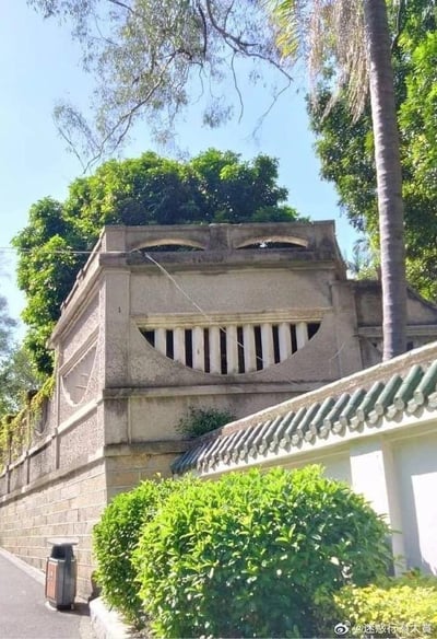 这张图片呈现了一处户外街景：路边的建筑墙体因独特的造型形成了有趣的视觉错位效果——墙体上方的两个弧形开口酷似眼睛，下方的弧形栅栏窗户如同咧开的嘴巴，搭配顶部生长的绿植，整体看起来像一张咧嘴笑的脸。画面下方有茂密的绿色灌木丛，左侧是道路和垃圾桶，背景有高大的树木，场景充满生活化的轻松感，这种自然形成的巧合让普通的建筑变得十分有趣。