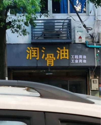 图片展示了一处街头店铺的招牌特写。招牌主体为深灰色底，上方用醒目的黄色大字书写“润骨油”，右侧配有白色小字“工程用油 工业用油”。背景中可见建筑物外墙、安装在金属格栅内的空调外机、带有红色警示三角形的电力设备，以及左侧伸出的绿色树枝。前景有一辆汽车的车顶部分遮挡视线。值得注意的是，招牌上的“润骨油”可能是“润滑油”的笔误，“滑”字误写为“骨”（意为骨头），形成了“滋润骨头的油”这一略带幽默的误解，与旁边“工程/工业用油”的专业用途形成反差。