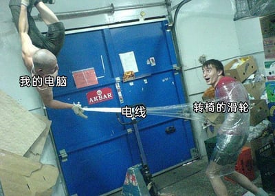 这是一张经过文字加注的真人搞笑梗图，画面设定在堆满纸箱的仓库环境中，背景是带有“AKBAR”标识的蓝色大门。左侧的光头男性未穿上衣，双手挂在上方管道上身体悬空，被标注为“我的电脑”，一只手抓着一根长条物体；右侧男性被透明保鲜膜缠绕身体并连在转椅上，表情惊讶、嘴巴大张，身体呈现出被拉扯的姿态，被标注为“转椅的滑轮”；两者之间的长条物体被标注为“电线”。这张图通过夸张的真人演绎，将日常使用电脑时的小场景拟人化：当电脑被挪动时，连接的电线会将坐在转椅上的人一同带着走，用具象的人物互动放大了这个尴尬又搞笑的日常瞬间，让观者产生共鸣。