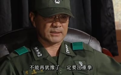 图片中是一位身穿绿色军装的中年男性军官，肩扛多颗星的军衔标志，头戴军帽，佩戴眼镜，正坐在黑色皮质椅子上。他表情严肃，背景为深色幕布，给人一种正式且庄重的感觉。画面下方有白色字幕：“不能再犹豫了 一定要出重拳”，显示出角色正在做出重要且果断的决定。