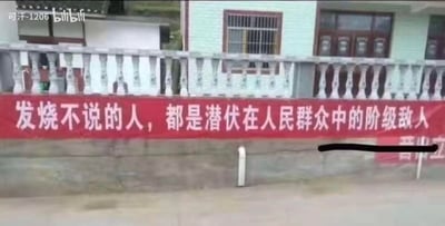 这是一张拍摄于乡村住宅外的实拍图，画面中住宅的石质栏杆下方悬挂着一条红色宣传横幅，横幅上用白色字体写着极具硬核风格的防疫标语：“发烧不说的人，都是潜伏在人民群众中的阶级敌人”。该标语借用带有年代感的表述方式，夸张地强调了发烧后主动上报的重要性，在防疫时期以直白强硬的语气起到宣传作用，同时也带有一定的调侃感。