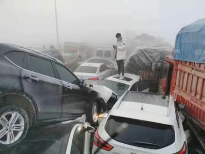 整张图拍摄于能见度极低的大雾天气下的道路区域，现场发生了大规模多车连环相撞的交通事故，大量私家车、货运卡车相互挤撞堆叠，多辆车辆出现变形损坏，事故拥堵范围很大。画面中央一名穿着白色羽绒服、黑色长裤、白色运动鞋的男子站在一辆受损白色轿车的车顶上，低头专注操作手机，在混乱的事故现场显得格外突出，周围满是相撞的车辆，远处浓雾中还能看到更多被困的车辆，整体场景混乱。