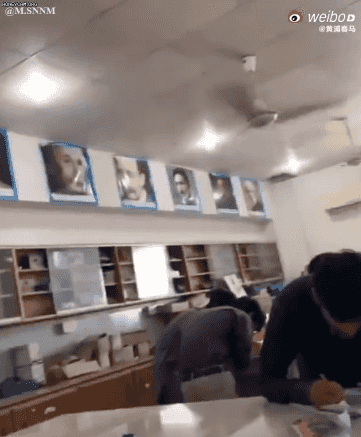 这是一则利用视觉错位效果的动态GIF图，场景为室内工作空间：墙面上的人物肖像照被悬挂在靠近天花板的位置，借助拍摄视角的巧妙设计，这些肖像看起来像是正低头凝视着下方桌前忙碌的人，营造出一种被严肃“大佬”紧盯的诙谐氛围，画面充满搞笑感，仿佛照片中的人在监督工作者，带来趣味十足的压迫感。