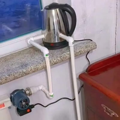 这张实拍图展示了一个极具脑洞的非规范DIY装置：普通不锈钢电热水壶被放在花岗岩材质的窗台上，两根白色PVC管道从壶身两侧接入，其中一根管道向下连接墙面上安装的加压水泵，另一根管道向下延伸，整体是将用来烧水的电热水壶违规改造成了热水循环供暖设备，改造方式十分粗糙，完全不符合用电和用水安全规范，充满荒诞搞笑的效果。