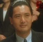 图片中是一位穿着深色西装、系着领带的男性，发型整齐，面部表情平静，正正面注视前方。背景中可见其他模糊的人影，推测是在正式场合或电影场景中。整体画面呈现出沉稳、正式的氛围，人物形象经典。