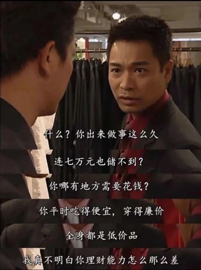 图片展示了电视剧《创世纪》中的经典场景，一位穿着西装的男性正在严肃地质问另一位男性同事的储蓄能力。背景中可以看到挂满西装的衣架，表明场景发生在服装店或西装店中。质问者表情严肃，语气严厉，连续发问对方为何工作多年仍无法储蓄到七万元，质疑其消费习惯和理财能力。