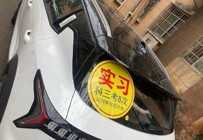 图片展示了一辆白色汽车的车尾部分，车尾贴有一个醒目的黄色圆形实习贴纸。贴纸上方用红色大字写着“实习”，下方黑色文字写着“科三考8次 比特斯拉还可怕”。这是车主通过幽默自嘲的方式，用夸张的说法（考驾照科三8次失败）来调侃自己的驾驶技术，同时借用“特斯拉”的品牌知名度制造反差笑点，暗示自己的驾驶状态比以加速快著称的特斯拉更令人“害怕”。