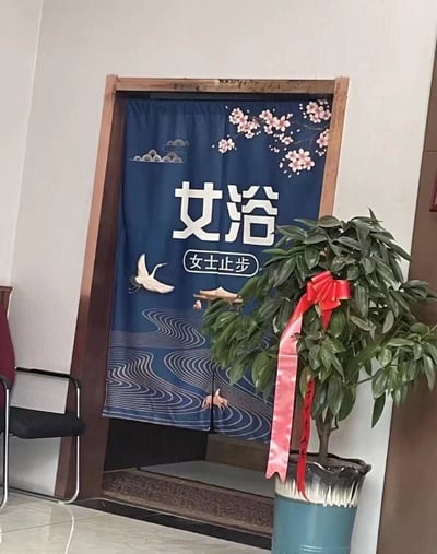 一张拍摄于室内的照片，展示了一个挂着蓝色门帘的门口。门帘上印有传统中国风图案，包括粉色梅花、仙鹤和波浪纹样。门帘正中央用白色大字写着“女浴”，下方却用白色小字标注“女士止步”，形成明显的矛盾和幽默效果。门帘右侧有一盆系着红色蝴蝶结的绿植，左侧能看到一把黑色椅子的部分轮廓。整个场景看起来像是在某个公共浴室或温泉场所。