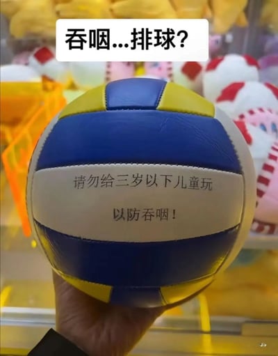 这张图片展示了抓娃娃机中的一个蓝黄白配色的排球，排球的白色区域印有“请勿给三岁以下儿童玩 以防吞咽！”的安全提示语。由于排球本身体积较大，三岁以下儿童完全不可能将其吞咽，这个不符合常识的提示显得十分荒谬。图片上方配文“吞咽...排球？”，以吐槽的语气放大了这个提示的不合理性，制造出搞笑的反差效果，引发观者对这种错误套用安全模板行为的调侃。