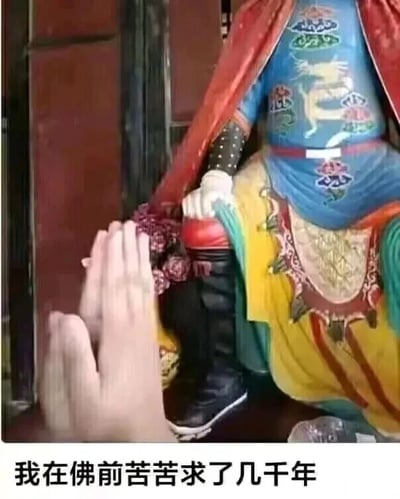 这是一张利用视觉错位效果创作的静态趣味图片。画面中一尊色彩艳丽的民俗神像（外观疑似关公）的手，与画面下方拍摄者的手，因拍摄角度的巧合，看起来像是紧紧握在一起。图片底部配有文字“我在佛前苦苦求了几千年”，这句文字出自经典流行歌曲《求佛》的歌词，结合视觉错位形成的“握手”效果，制造出一种仿佛拍摄者真的在和神像互动、呼应歌词里“求佛”意境的搞笑反差感，整体风格诙谐幽默，适合用来调侃自己祈求愿望的状态，或是单纯用来活跃社交氛围。