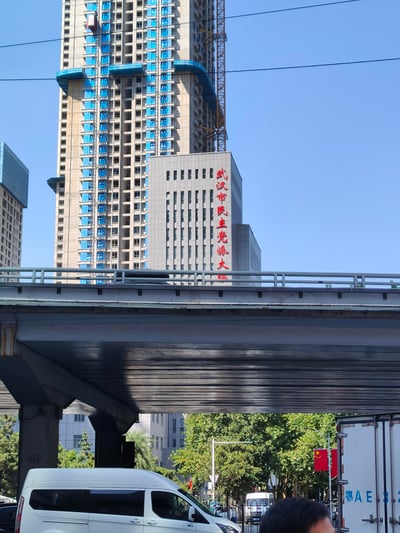 这是一张晴朗白天在武汉街头拍摄的城市实景照片，画面上方是带有施工塔吊的在建超高层住宅楼，楼体为未完工的混凝土结构并带有蓝色防护设施；中景是灰色外立面的武汉市民主党派大楼，红色竖排字样清晰可见；前景是城市高架桥的底部，桥下道路上有白色厢式车辆，路边还悬挂着中国国旗，整体展现了武汉城市中新旧建筑并存的日常风貌。