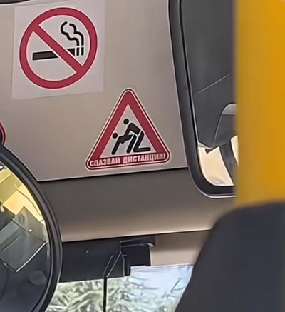 车辆内部车门面板的特写照片，展示了两个警示贴纸。上方为标准的红色圆形禁止吸烟标识，中间有香烟图案和斜杠；下方为红色边框三角形警告标识，内部有两个人形图案，下方配有西里尔文字"СОХРАНЯЙТЕ ДИСТАНЦИЮ"（保持距离）。整体画面清晰，聚焦于警示标识的细节展示。