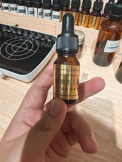 一只手拿着一瓶棕色滴管款精油瓶，瓶身金色标签印有"特朗普（香） 高定沙龙香"字样，背景是手工调香的工作台，台面上摆放着多款调香原料瓶（可见"康乃馨""东方香""樟脑""苦咖啡""尤加利"等标签）、标注"陈化剂"的容器，还有带瞄准线的电子秤设备，整体呈现手工定制香氛的制作场景，无谐音梗或暗示内容，仅为一款以"特朗普"命名的定制沙龙香产品实拍。