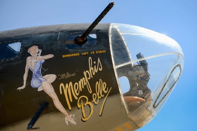 这张照片展示了二战著名轰炸机"孟菲斯美女号"(Memphis Belle)的机头特写。照片采用仰拍角度，清晰呈现了飞机标志性的机头艺术——一位身着蓝色紧身衣的女性侧身像，旁边印有金色字体的机名"Memphis Belle"和"BOMBARDIER CAPT. VB EVANS"字样。机头前部装有透明座舱罩，内部可见部分机械结构，侧面还装有一挺机关枪。背景是纯净的蓝天，突出展现了这架传奇轰炸机的经典设计和历史质感。