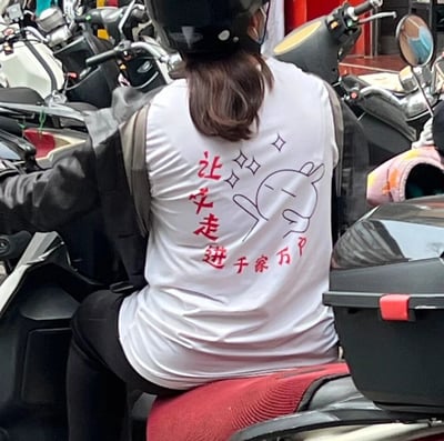 这是一张街头实拍的静态图，画面主体是一位戴着黑色头盔的女性，她坐在电动车后座，内搭的白色T恤背部印有红色标语“让牛走进千家万户”，旁边搭配了一个比耶的卡通兔子简笔画。周围停放着多辆电动车，场景看起来是一处日常的电动车停放点，T恤上的标语结合卡通图案，风格接地气且带有趣味感。