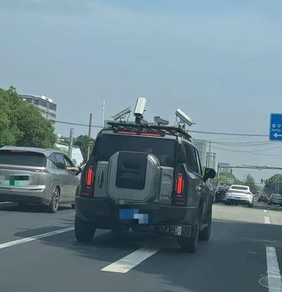 一张在城市道路上拍摄的照片，画面中心是一辆黑色SUV，其车顶安装了多个朝向不同方向的监控摄像头，车辆正在正常行驶。周围有其他社会车辆，背景可见树木、建筑物及一个蓝色的道路指示牌，上面写有“华宁”字样及向左的箭头。整体场景像是日常交通环境，因SUV顶部的特殊摄像头配置显得较为特别，可能引发对车辆用途的猜测。