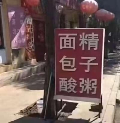 照片展示了一条具有传统风貌的街道，背景可见挂着红色灯笼的古建筑，墙面有红色装饰元素。街道旁立着一块红色立式招牌，牌面用白色字体纵向书写“面精”“包子”“酸粥”三组文字，推测为当地特色小吃的宣传标识。画面光线明亮，呈现出日常街头的真实场景，充满生活气息。