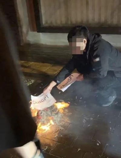 图中一名面部被打码的男子蹲在室外的砖石地面上，正将写有“ChatGPT”字样的纸张投入面前的火堆中焚烧，地面上的火焰正在燃烧。该场景模仿了传统焚烧祭祀的行为，以一种夸张戏谑的网络玩梗方式，表达对ChatGPT这款热门AI聊天工具的调侃或情绪宣泄，是网友针对AI相关话题创作的搞笑内容。