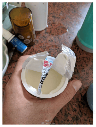 这是一张原始拍摄的生活照片，画面主体是一只手握着一个打开的杯装酸奶。酸奶的盖子被撕开后，盖子上的白色塑料提手与杯内放置的吸管包装在视觉上恰好重合，看起来仿佛这个带有文字的吸管包装是从提手中延伸出来的，形成了有趣的视觉错位效果。背景是带有红棕色纹理的桌面，上面还摆放着棕色带白点的容器、蓝色小瓶、白色容器等物品，图片底部带有“日品”和“ADnmb.com”的水印。